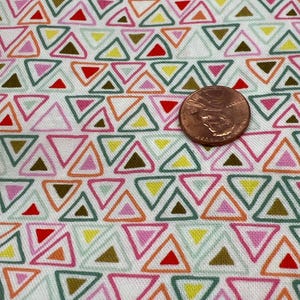 Puede incluir: Tela blanca con un patrón repetido de triángulos coloridos. Los triángulos están delineados en verde, rosa, naranja y marrón. Una moneda de color cobre se coloca sobre la tela.