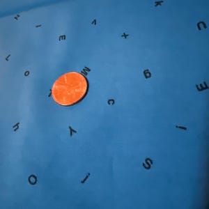 Puede incluir: Primer plano de una superficie azul con letras impresas en ella. Un centavo de cobre se coloca sobre la superficie.