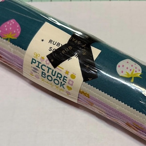 Könnte beinhalten: Eine Rolle aus Stoffquadraten, eingewickelt in Klarsichtfolie mit einer Papierbanderole, beschriftet mit Ruby Star Society Picture Book von Kimberly Kight. Der Stoff zeigt Muster aus rosa und weißen Erdbeeren auf dunkelblaugrünem Grund.