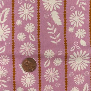 May include: Pink fabric featuring a repeating pattern of white daisies, leaves, and small floral elements. Vertical brown zig-zag stripes separate the floral designs. A penny is included for scale.