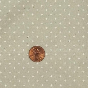 Puede incluir: Una tela de color gris claro con un patrón repetitivo de pequeñas cruces blancas. Una moneda de cobre de los Estados Unidos está colocada sobre la tela para proporcionar una referencia de escala, midiendo aproximadamente 1,9 cm de diámetro.