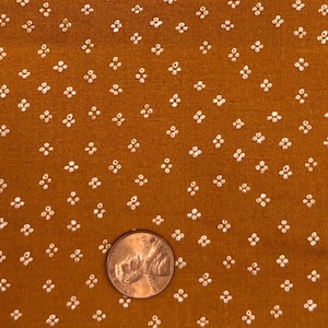 May include: Close-up of a fabric with a repeating pattern of small white floral-like shapes on a mustard-colored background. A U.S. penny is included for scale. The fabric has a textured appearance.