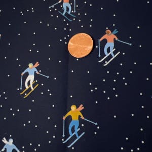 Puede incluir: Tela azul oscuro con un patrón repetido de esquiadores con trajes coloridos y puntos blancos que se asemejan a copos de nieve. Una moneda está en el centro de la tela. Los esquiadores llevan trajes azules, naranjas y amarillos.