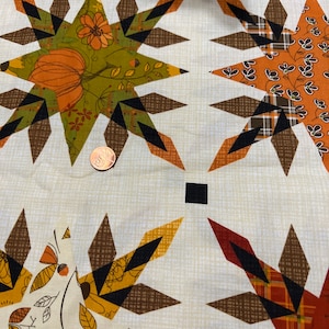 Puede incluir: Una tela beige con un patrón repetido de formas de estrellas marrones, naranjas y negras. Las estrellas están formadas por formas geométricas más pequeñas y tienen un diseño de patchwork. La tela tiene una textura sutil.