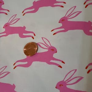May include: White fabric with a repeating pattern of pink bunnies hopping. The bunnies have red accents on their feet and ears.