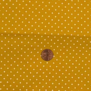 Puede incluir: Una tela de color amarillo mostaza con un patrón repetitivo de pequeños signos de más blancos. Una moneda de color cobre está colocada en el centro de la tela para proporcionar una escala del tamaño del patrón de signos de más impreso.
