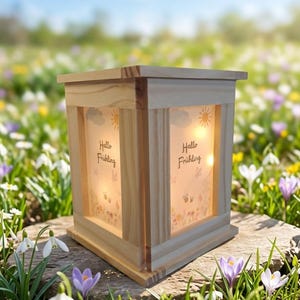 Könnte beinhalten: Eine Holzlaterne mit dem Text "Hallo Frühling" auf den Frontplatten. Die Platten haben ein Blumendesign mit Schmetterlingen, Wolken und Sonnen. Die Laterne ist von innen beleuchtet und steht auf einer Steinoberfläche, umgeben von Blumen.