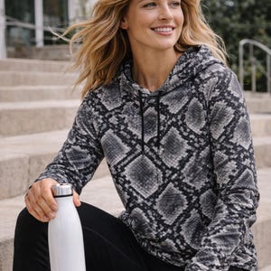 May include: A long-sleeve hooded sweatshirt featuring a grey and black snakeskin pattern with a black drawstring, paired with solid black leggings and a white stainless steel water bottle with a silver cap.
