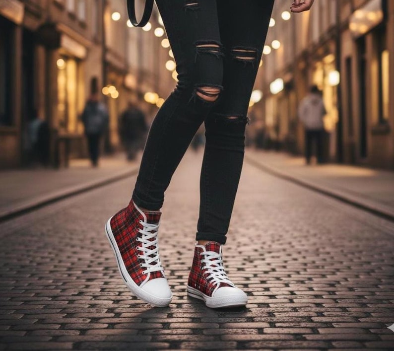 Pu&ograve; includere: Sneakers alte con motivo tartan rosso e nero, lacci bianchi e punte in gomma bianca, indossate con jeans skinny neri strappati con tagli orizzontali sulle ginocchia, su uno sfondo urbano sfocato.