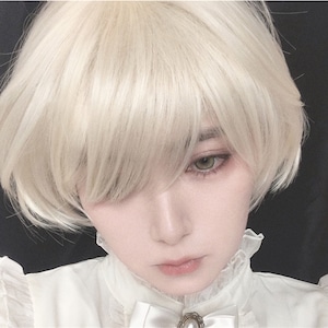May include: A person with pale skin and light green eyes wearing a short, light blonde wig. The person is wearing a white blouse with a ruffled collar and a pearl brooch.