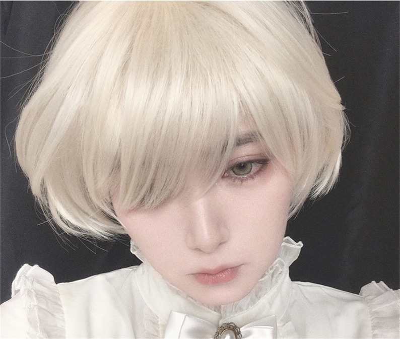 May include: A person with pale skin and light green eyes wearing a short, light blonde wig. The person is wearing a white blouse with a ruffled collar and a pearl brooch.