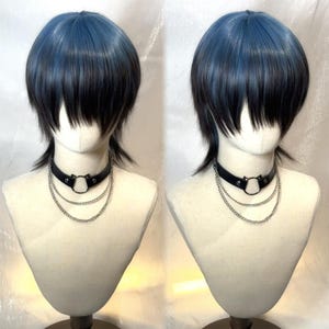 Puede incluir: Peluca bicolor con capas azules y negras, peinada con un corte corto y en capas. La peluca se exhibe en una cabeza de maniquí, combinada con un collar de gargantilla de cuero negro con detalles de cadena plateada.