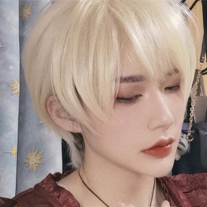 May include: A close-up of a person with short, light blonde hair styled with a side-swept fringe. The person has fair skin, with visible eye makeup and red lipstick. A dark necklace is visible.