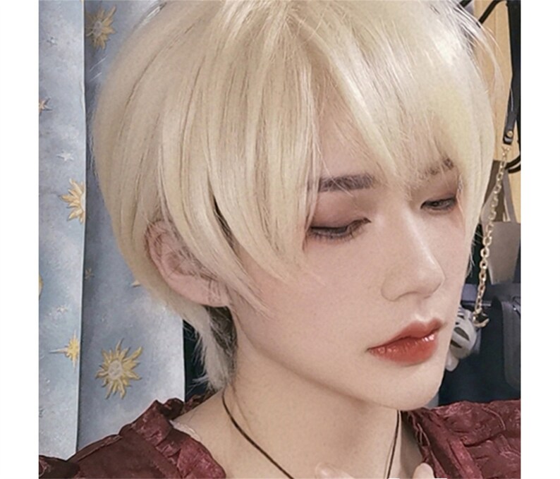 May include: A close-up of a person with short, light blonde hair styled with a side-swept fringe. The person has fair skin, with visible eye makeup and red lipstick. A dark necklace is visible.