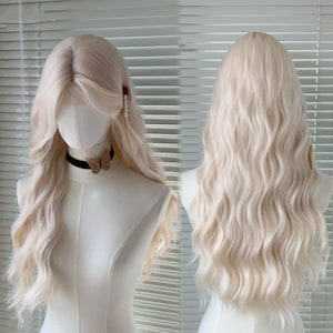 Puede incluir: Peluca rubia clara y ondulada exhibida en un maniquí. La peluca presenta una raya lateral, capas que enmarcan la cara y un estilo voluminoso. El maniquí lleva un collar negro con un colgante de mariposa.