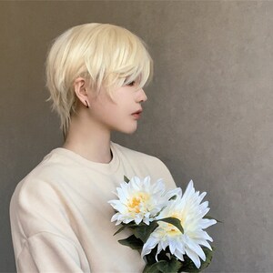May include: A person wearing a light beige sweatshirt and a short, layered, light blonde wig. The person is holding two white and yellow artificial flowers with green leaves.