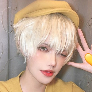 May include: A person with short, light blonde hair and wearing a yellow hat and sweater. The person is winking and holding up a hand with a yellow heart emoji. The person has light skin and is wearing makeup.