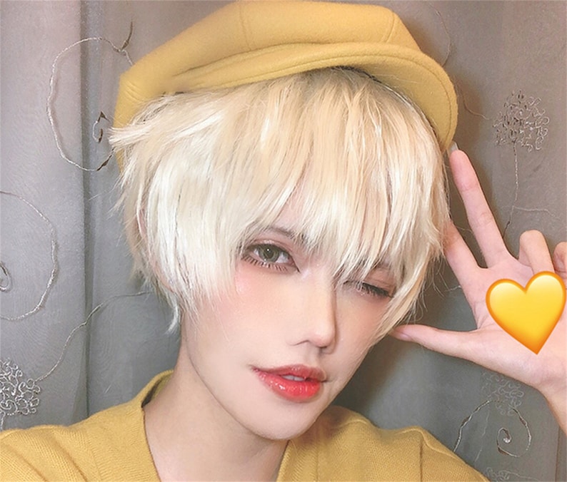May include: A person with short, light blonde hair and wearing a yellow hat and sweater. The person is winking and holding up a hand with a yellow heart emoji. The person has light skin and is wearing makeup.