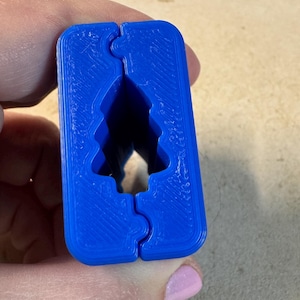 Könnte beinhalten: Ein hellblaues, 3D-gedrucktes Objekt mit einem Weihnachtbaum-Ausschnitt. Das Objekt ist rechteckig mit ineinandergreifenden Teilen. Die Baumform ist als negativer Raum im blauen Kunststoff sichtbar.
