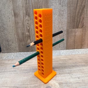 Könnte beinhalten: Ein leuchtend orangefarbener, vertikaler Stiftehalter mit quadratischem Sockel und zwei Reihen runder Löcher. An der Seite befinden sich eingeprägte Zahlenmarkierungen von 1,5 bis 15,5. Zwei Stifte, einer schwarz und einer dunkelgrün, stecken in den Löchern.