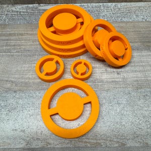 Könnte beinhalten: Orangefarbene, 3D-gedruckte Objekte in verschiedenen Größen. Einige sind gestapelt, mit den Maßen 8cm, 9cm und 10cm. Die Objekte haben ein kreisförmiges Design mit einer zentralen Scheibe und einem umlaufenden Ring.