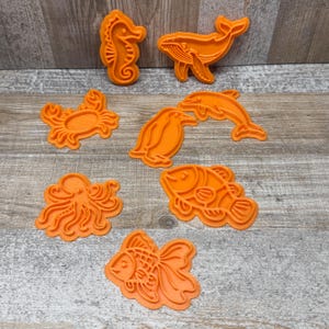 Könnte beinhalten: Orange Ausstecher in Form von Meerestieren, darunter Seepferdchen, Wal, Krabbe, Pinguin, Delfin, Oktopus und Fisch. Ideal zum Backen von Keksen oder für Bastelprojekte.