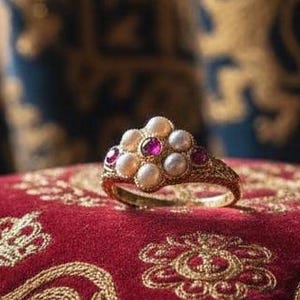 May include: A gold ring with pearls and red gemstones rests on a red velvet pillow with gold embroidery. The ring is the focal point, showcasing its intricate design and the contrast of colors.