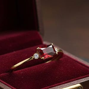 Puede incluir: Un anillo de oro con una piedra preciosa rectangular de color rojo intenso y dos piedras preciosas blancas más pequeñas. El anillo se muestra en un joyero de madera abierto con interior de terciopelo rojo. La caja tiene un cierre dorado.