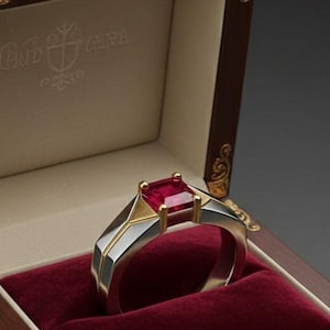 Puede incluir: Un anillo de rubí en una caja de joyería de madera. El anillo presenta una gema roja de corte cuadrado engastada en una banda plateada con detalles dorados. La caja es marrón oscuro con filigrana dorada y un interior de terciopelo rojo.