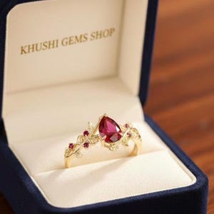 Peut inclure: Une bague en or avec une pierre précieuse rouge foncé en forme de poire est présentée dans un écrin de bijoux en velours bleu. La bague présente un motif floral avec de petites pierres rouges. La boîte ouverte affiche le texte "KHUSHI GEMS SHOP".
