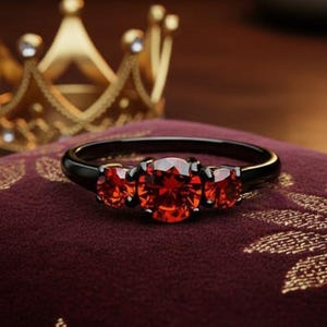 May include: A black ring with three red gemstones, set on a burgundy cushion with a gold floral pattern. A gold crown is in the background. The ring's gemstones are a vibrant red color.