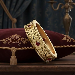 May include: A gold-colored bracelet with intricate filigree and red gemstone accents rests on a burgundy velvet pillow with gold embroidery. The bracelet's design features repeating patterns and square-cut gemstones.