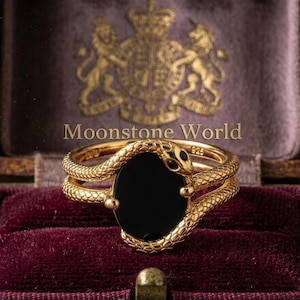 Puede incluir: Un anillo de tono dorado con un motivo de serpiente texturizada que envuelve una piedra negra lisa de forma ovalada. La banda está marcada con 925, indicando plata de ley. El texto Moonstone World es visible encima del anillo.