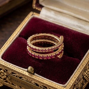 May include: Two gold rings, each set with a continuous line of red gemstones, displayed in a vintage jewellery box with a burgundy velvet lining. The rings are stacked, highlighting the detailed setting and stones.