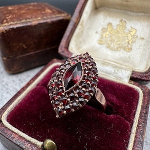 Könnte beinhalten: Ein marquiseförmiger Ring mit einem zentralen, länglichen, dunkelroten Granatstein, der von mehreren kleineren facettierten Granatsteinen in einer Metallschiene umgeben ist. Der Ring liegt in einer dunkelbraunen Schmuckschatulle.