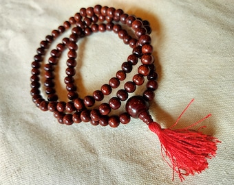 May include: A strand of prayer beads made of dark brown wooden beads. The beads are strung together and have a large bead at the end with a red tassel. The beads are resting on a light beige fabric.