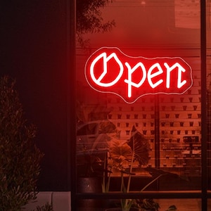 May include: A red neon "Open" sign in a window. The sign has a white outline and is in a stylized font. The sign is in front of a restaurant or shop. A potted plant is in the foreground.