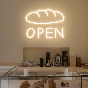 May include: A neon sign with a bread loaf shape above the word "OPEN" illuminates a bakery. Display cases showcase pastries, including croissants and other baked goods. Coffee machines and serving dishes are visible.