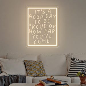 May include: A rectangular, illuminated sign with the text "IT'S A GOOD DAY TO BE PROUD OF HOW FAR YOU'VE COME" in a handwritten style. The sign is mounted on a neutral-colored wall above a white sofa with decorative pillows and a coffee table.