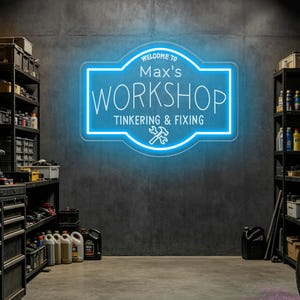 May include: A bright blue neon sign mounted on a dark gray wall. The sign features a rectangular shape with rounded corners and displays the text "WELCOME TO Max's WORKSHOP TINKERING & FIXING" along with a graphic icon of a crossed hammer and wrench.