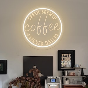 Może przedstawiać: Biały neonowy znak z napisem "Fresh Brewed Coffee Served Daily" w kształcie koła. Znak wisi na białej ścianie nad drewnianym szafką z ekspresem do kawy i innymi przedmiotami.