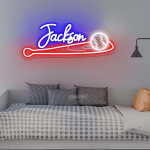 May include: A neon sign in the shape of a baseball bat and ball, with the name "Jackson" in blue script. The bat is red, and the baseball is white. The sign is mounted on a light gray wall above a bed with gray and white bedding.