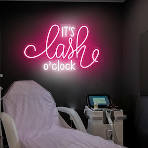May include: A pink neon sign saying "It's lash o'clock" is mounted on a dark grey wall. A light pink treatment chair is below the sign. A medical device and a towel rack are also visible in the room.