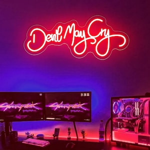 May include: A neon sign displaying the words "Devil May Cry" in a cursive script, illuminated in white. The sign is mounted on a wall above a gaming setup with dual monitors, a keyboard, and a computer tower.