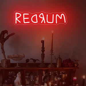 Puede incluir: Un letrero de neón rojo que dice "REDRUM" cuelga sobre una estantería de madera llena de velas, objetos decorativos y una pequeña estatua de gato. La escena está tenuemente iluminada, creando una atmósfera espeluznante. El letrero es el punto focal.