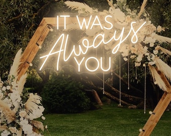 May include: A wooden hexagonal wedding arch decorated with white flowers and pampas grass. The neon sign reads "IT WAS Always YOU". The arch is set on a grassy lawn with trees in the background.