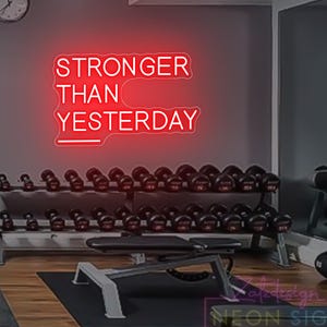 Könnte beinhalten: Rotes Neon-Schild mit der Aufschrift "STRONGER THAN YESTERDAY" in einem Fitnessstudio. Das Bild zeigt Hanteln auf einem Ständer, eine Hantelbank und eine Uhr. Ideal für ein Fitnessstudio zu Hause oder ein Fitnessstudio.