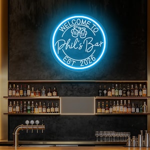 May include: A circular blue neon sign mounted on a dark wall, featuring the text "WELCOME TO Phil's Bar EST 2026" with a graphic of two clinking beer mugs in the centre. The sign emits a bright glow, illuminating the surrounding bar area.