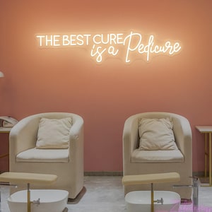Könnte beinhalten: Ein Leuchtschild mit der Aufschrift "THE BEST CURE is a Pedicure" hängt an einer rosa Wand. Darunter stehen zwei beige Sessel mit Kissen. Jeder Stuhl hat ein Fußbad und einen kleinen Tisch.