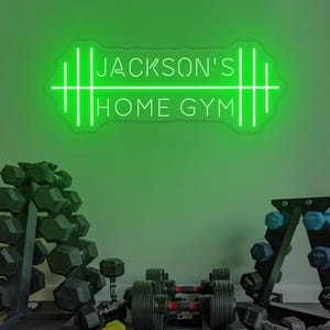 以下が含まれることがあります： 「Jackson's Home Gym」と書かれた緑色のネオンサインが、ダンベルとウェイトラックのコレクションの上に吊り下げられています。ダンベルは黒と黄色で、ウェイトラックは黒です。背景は単色の緑色です。
